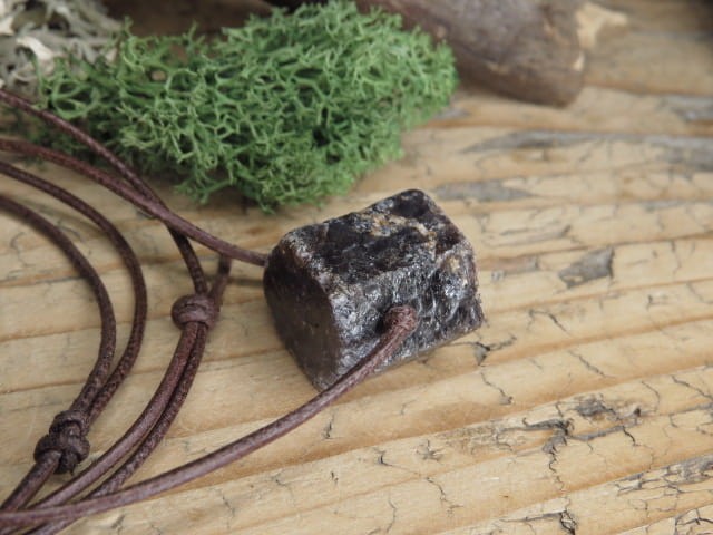 Korund na rzemieniu. Kryształ korundu - kamień naturalny - pojedynczy kryształ. Kamień nie jest szlifowany. Kamień przewiercony, zawieszony na skórzanym rzemyku w kolorze brązowym.