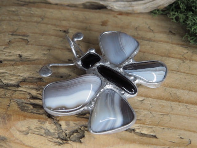 Wyjątkowy, ręcznie wykonany wisior artystyczny w kształcie motylka, stworzony z naturalnych kamieni: agatu i onyksu (środek). Całość została oprawiona w miedź i cynę, co nadaje kompozycji szlachetnego charakteru i surowego uroku.