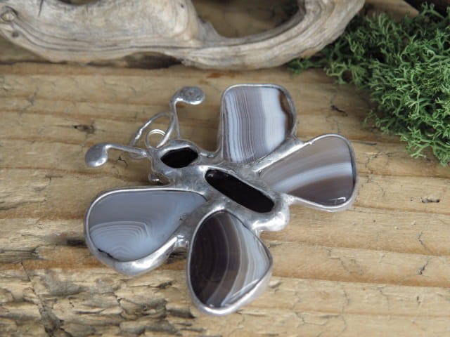 Wyjątkowy, ręcznie wykonany wisior artystyczny w kształcie motylka, stworzony z naturalnych kamieni: agatu i onyksu (środek). Całość została oprawiona w miedź i cynę, co nadaje kompozycji szlachetnego charakteru i surowego uroku.