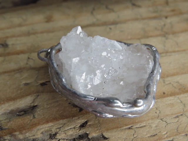 Ręcznie wykonany wisior z naturalną szczotką krystaliczną kryształu górskiego. Liczne, błyszczące kryształki tworzą efektowną, trójwymiarową strukturę, która pięknie odbija światło i podkreśla naturalne piękno minerału.